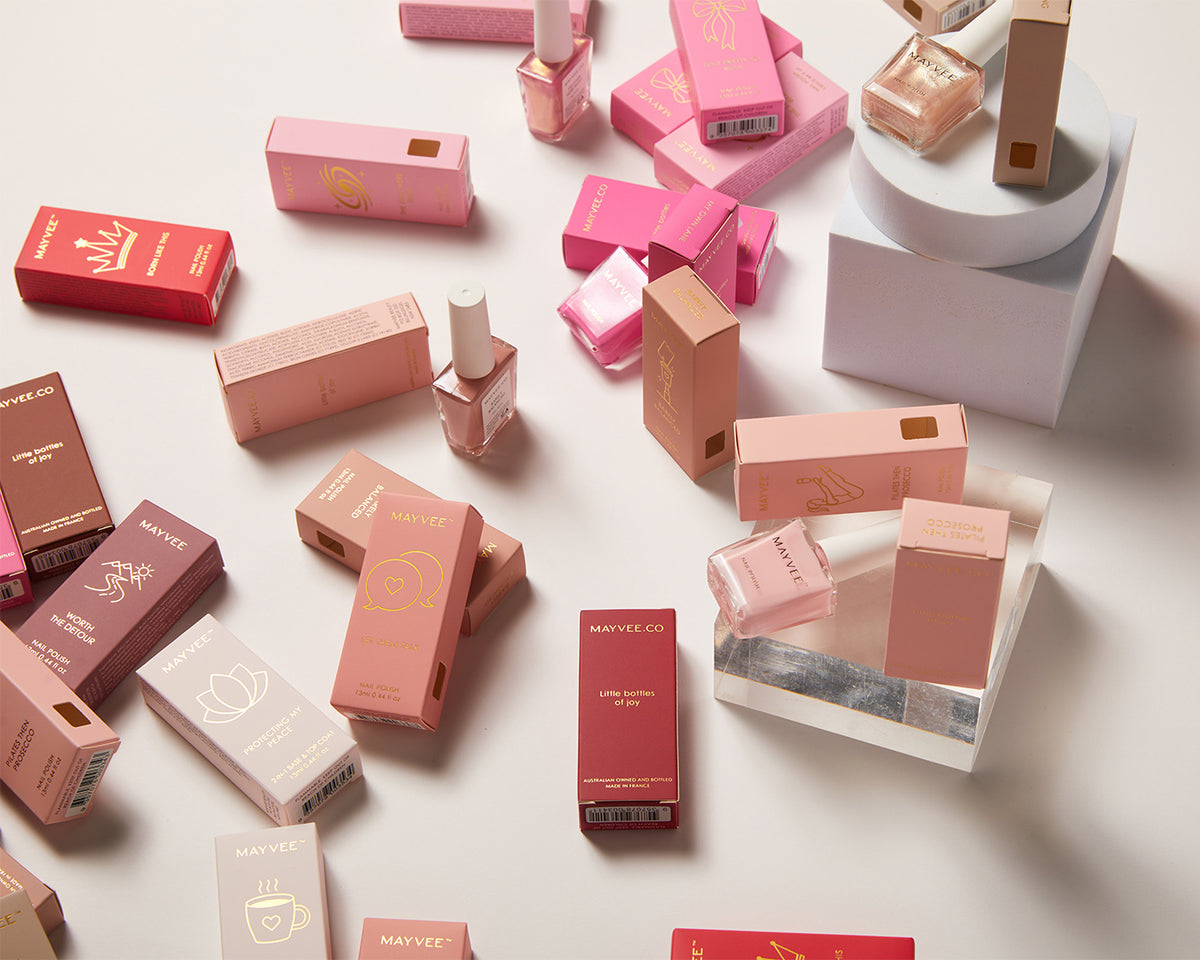 An assortment of Maryvee cosmetics, including lipsticks, nail polishes, and boxes, arranged in a styled flat-lay on a white surface with a white pedestal.